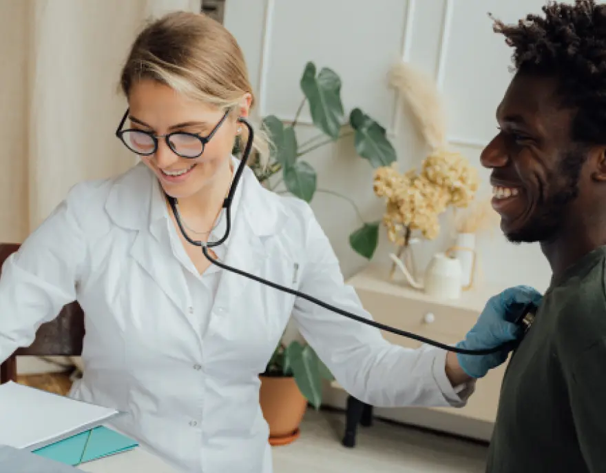 A doctor smiles as she listens to a laughing patient’s chest with a stethoscope.