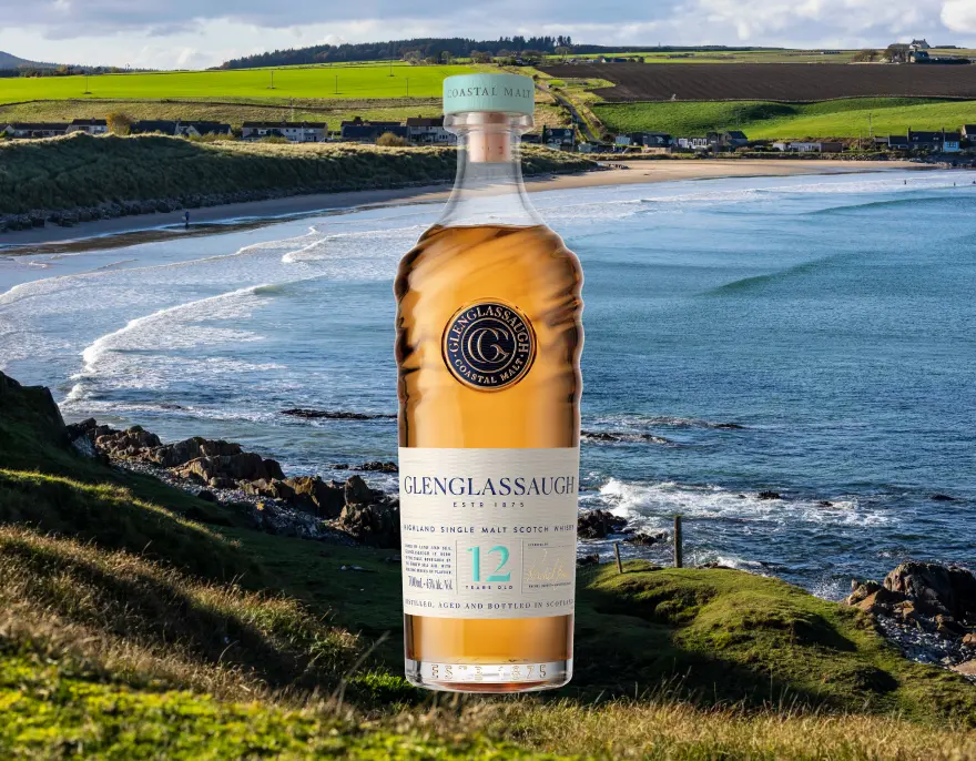 A bottle of Glenglassaugh 12 on a grassy clifftop above a sandy bay.