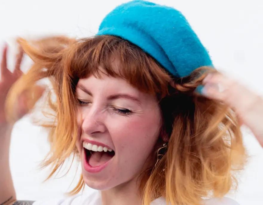 A laughing young woman in a blue beret shakes out her red hair.