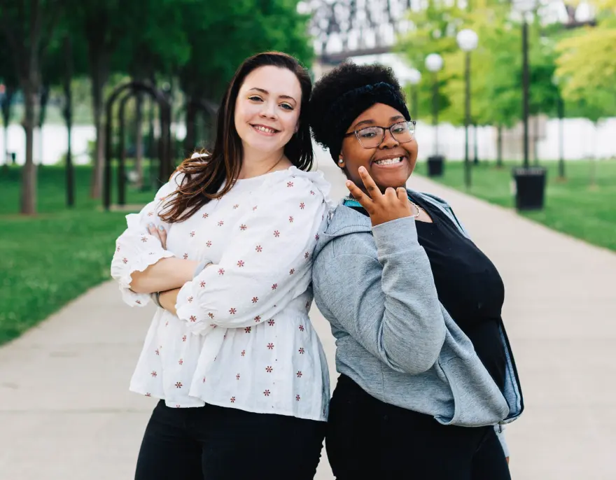 Two smiling women stand back to back on a tree-lined riverside path.