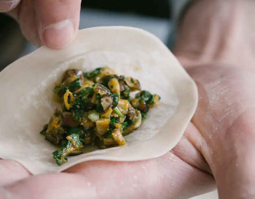 Hands fold a dumpling wrapper around a filling of greens and mushrooms.