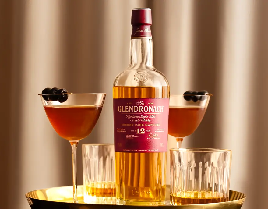 A bottle of The Glendronach 12 on a gold tray between two cocktails and glasses of whisky.