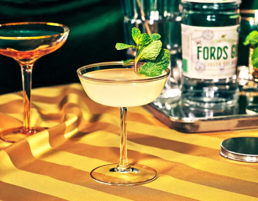 A gin cocktail garnished with mint beside a bottle of Fords Gin on a gold striped cloth.